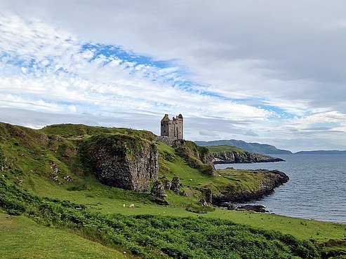 Das Foto zeigt Kerrera Castle in Schottland, mit stimmungsvollem wolkigen Himmel, auf saftig-grüner Wiese, am rechten Bildrand das Meer.